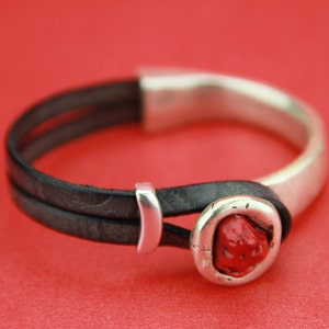 May include: A black leather bracelet with a silver band and a red stone accent.