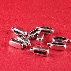 May include: A collection of silver-colored metal cord ends, oval-shaped with a small loop at one end, scattered on a red background. These findings are likely used in jewelry making or crafting projects.