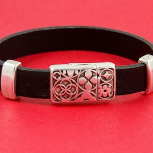 May include: Black leather bracelet with a silver clasp. The clasp features an ornate floral design. The bracelet is approximately 0.5 inches wide and has silver end caps. The background is a solid red.