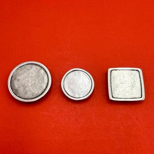 May include: Three silver-toned metal components with recessed centers. Two of the pieces are circular with smooth, raised rims, while the third piece is square with rounded corners and a matching raised border, all featuring a matte, textured finish.