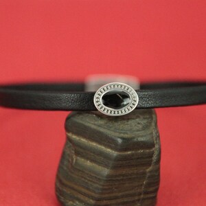 May include: A black leather bracelet with a silver oval charm featuring a black stone in the center.