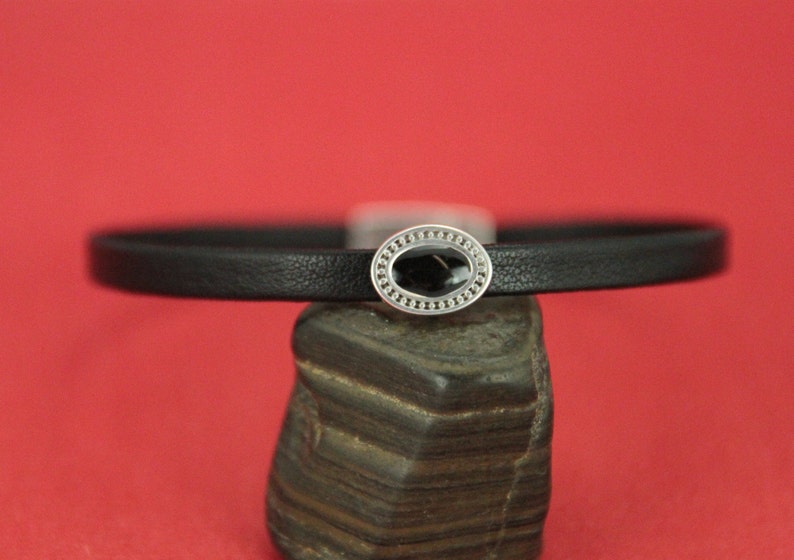 May include: A black leather bracelet with a silver oval charm featuring a black stone in the center.
