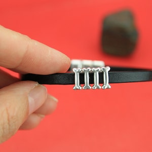 May include: A black leather bracelet with silver bone charms. The bracelet has four bone charms in a row.