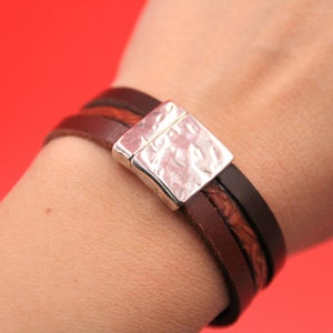 May include: A brown leather bracelet with a silver square clasp. The bracelet has three strands of leather in varying shades of brown. The clasp has a hammered texture. The bracelet is shown on a wrist against a red background.