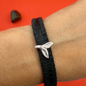 May include: Black leather bracelet featuring a silver-colored whale tail charm. The bracelet is a simple band with a textured surface. The charm is detailed with a textured finish, adding a touch of nautical flair.