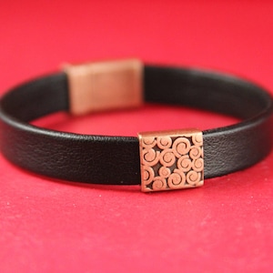 May include: A black leather bracelet with a copper-coloured clasp featuring a swirling pattern.