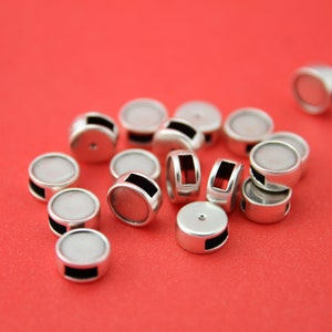 May include: A collection of silver-colored, cylindrical beads with a flat, white top and a black rectangular accent. The beads are scattered on a bright red background, creating a visually appealing contrast. The beads appear to be jewelry components.