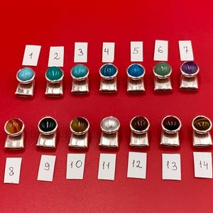May include: Fourteen silver charms with different colored gemstones. The charms are numbered 1 through 14.