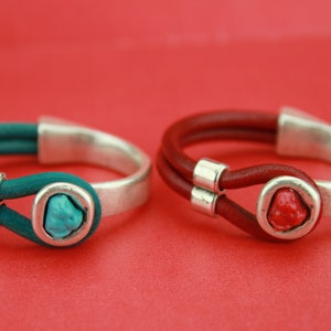 May include: Two leather bracelets with silver-tone metal accents and gemstone details. One bracelet features teal leather and a turquoise stone, while the other has red leather and a red stone. The bracelets are set against a red background.