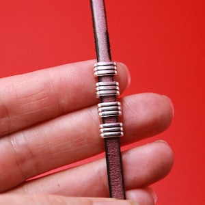May include: A close-up of a brown leather cord with silver metal beads. The beads are cylindrical and have a grooved design.