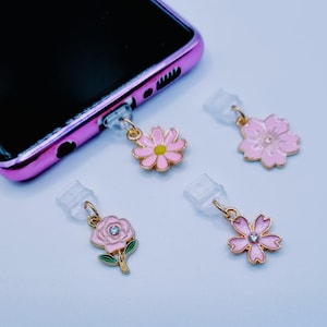 Peut inclure: Quatre breloques de téléphone roses à motifs floraux avec des accents dorés. Les breloques comprennent une marguerite, une rose et deux motifs de fleurs différents, chacun avec une prise transparente pour le port de charge du téléphone. Le téléphone est rose.