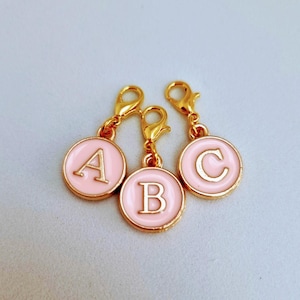 May include: Three gold-coloured letter charms with pink enamel. The charms are in the shape of circles with the letters A, B, and C in white.