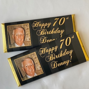 May include: Two black and gold chocolate bar wrappers with a photo of a man and the text "Happy 70th Birthday Denny!"