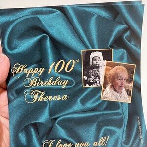 Puede incluir: Una tarjeta de sat&eacute;n azul turquesa con letras doradas que dice "Happy 100th Birthday Theresa I love you all!" Hay dos fotos en la tarjeta, una de una ni&ntilde;a peque&ntilde;a y otra de una mujer mayor.