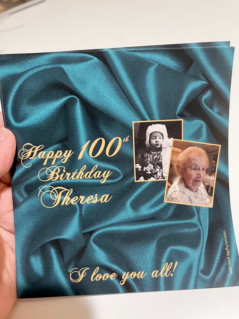 Puede incluir: Una tarjeta de sat&eacute;n azul turquesa con letras doradas que dice "Happy 100th Birthday Theresa I love you all!" Hay dos fotos en la tarjeta, una de una ni&ntilde;a peque&ntilde;a y otra de una mujer mayor.