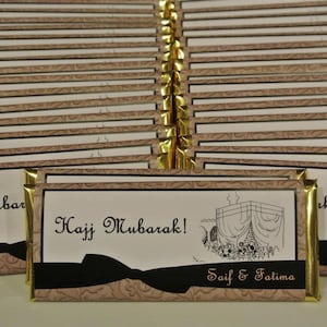 Puede incluir: Una pila de envoltorios de barras de chocolate con bordes dorados con el texto "Hajj Mubarak!" y "Saif & Fatima" impreso en ellos. Los envoltorios presentan una ilustración en blanco y negro de una mezquita.