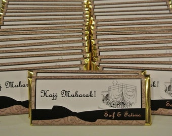 May include: A stack of gold-trimmed chocolate bar wrappers with the text "Hajj Mubarak!" and "Saif & Fatima" printed on them. The wrappers feature a black and white illustration of a mosque.