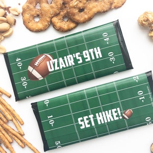 Puede incluir: Dos envoltorios de barra de chocolate verde y blanco con temática de fútbol con el texto "Uzair's 9th" y "Set Hike!" impreso en ellos. Los envoltorios presentan un diseño de campo de fútbol con líneas de yarda y un balón de fútbol marrón.