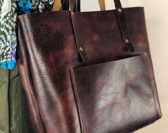 May include: A large, brown leather tote bag with a front pocket. The bag has a brown leather strap and a top zipper closure.