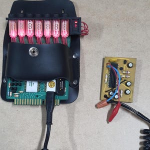 May include: A black leather case contains a circuit board and seven glowing red digital display tubes, each displaying the number 8. A small circuit board with wires and clips is next to the case. A black cable connects the case to a power source.