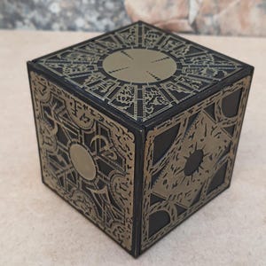 Może przedstawiać: Czarna i złota metalowa skrzynka z zagadką z misternymi wzorami na wszystkich stronach. Skrzynka ma okrągły otwór na górze.