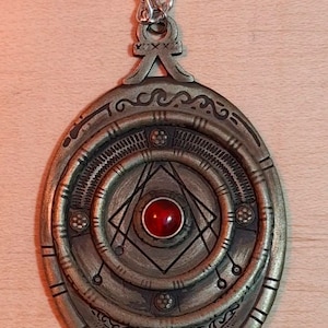 May include: A silver chain necklace with a large, oval pendant. The pendant is made of metal and has a circular design with a red gemstone in the centre. The pendant is surrounded by intricate details and patterns.