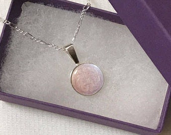 May include: A silver chain necklace with a round pendant featuring a pink and white swirl design. The necklace is displayed in a purple box with white lining.