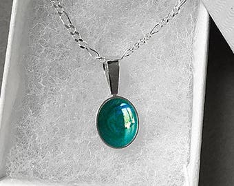 May include: A silver necklace with a teal-colored oval pendant rests inside a white gift box. The pendant has a glossy finish, and the chain is a delicate link style. The box is open, revealing the necklace against a backdrop of white fluff.