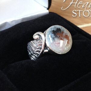 May include: A silver ring with a white, brown, and black stone set in a round bezel. The ring features a detailed wing design on the band. The ring is in a black velvet ring box. Heaven Stones is printed on the box.