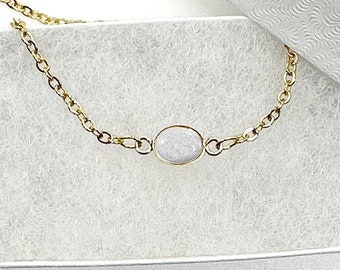 May include: A gold chain necklace with a single oval-shaped white stone pendant. The necklace is displayed in a white box with a textured lining.