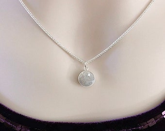 May include: A silver chain necklace with a round pendant containing a gray memorial stone. The pendant is attached to the chain with a silver bail. The necklace is on a model wearing a dark purple shirt. The text "HEAVEN STONES® from Memorial Silver" is visible above the necklace.