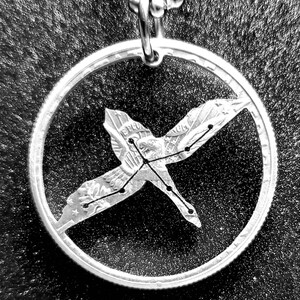 May include: A silver quarter coin necklace with a cutout of the constellation Cygnus, the swan, in the center. The coin is on a black background.