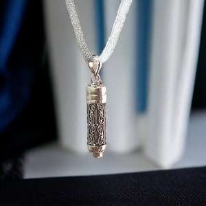 Può includere: Una collana con pendente color argento con un ciondolo cilindrico ornato da dettagli intricati in filigrana. Il ciondolo è appeso a una delicata catena d'argento intrecciata. Il pendente ha un design a vite. Lo sfondo è sfocato, con toni blu e bianchi.