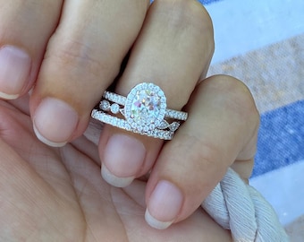 May include: A close-up of a hand wearing a silver oval-shaped engagement ring with a halo setting and two smaller diamond bands.