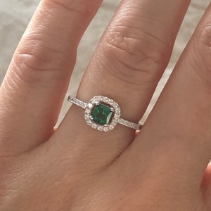 May include: A silver ring with a square emerald green gemstone surrounded by a halo of smaller clear gemstones.