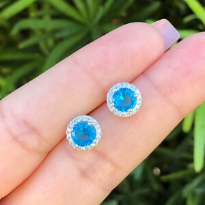 May include: A pair of silver stud earrings with a blue gemstone surrounded by a halo of smaller white gemstones.