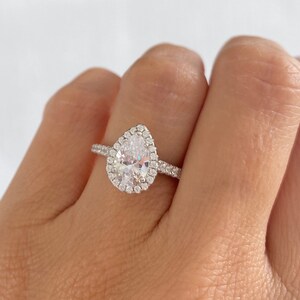 May include: A pear-shaped engagement ring with a halo setting. The ring is made of silver and has a clear center stone.