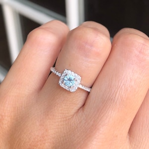 Pode incluir: Um anel de prata com uma pedra preciosa azul de forma quadrada cercada por pequenas pedras brancas.