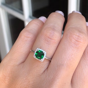 Puede incluir: Un anillo de plata con una piedra preciosa cuadrada verde esmeralda rodeada por un halo de piedras preciosas m&aacute;s peque&ntilde;as y transparentes.