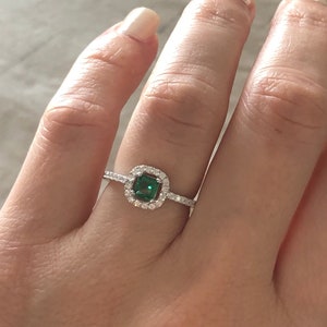 May include: A silver ring with a square emerald green gemstone surrounded by smaller clear gemstones.