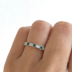 May include: A silver ring featuring a repeating pattern of square emerald-green stones and round clear stones. The ring is set with small, clear accent stones. The ring is shown on a person's finger.