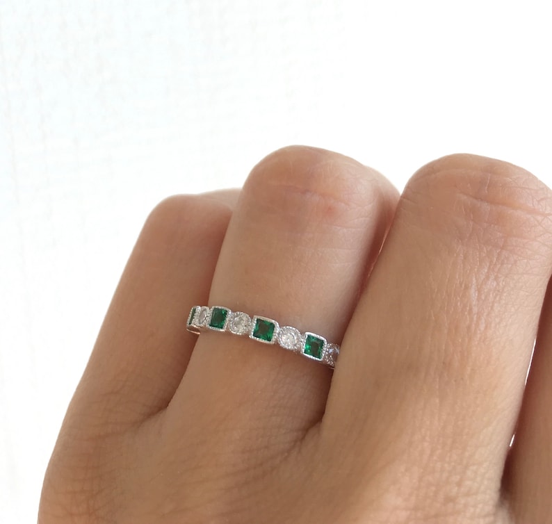 May include: A silver ring featuring a repeating pattern of square emerald-green stones and round clear stones. The ring is set with small, clear accent stones. The ring is shown on a person's finger.