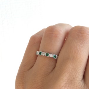May include: A silver ring with a repeating pattern of square emerald-green stones and round clear stones. The ring is set with small, round accent stones between the larger stones. The ring is on a finger.