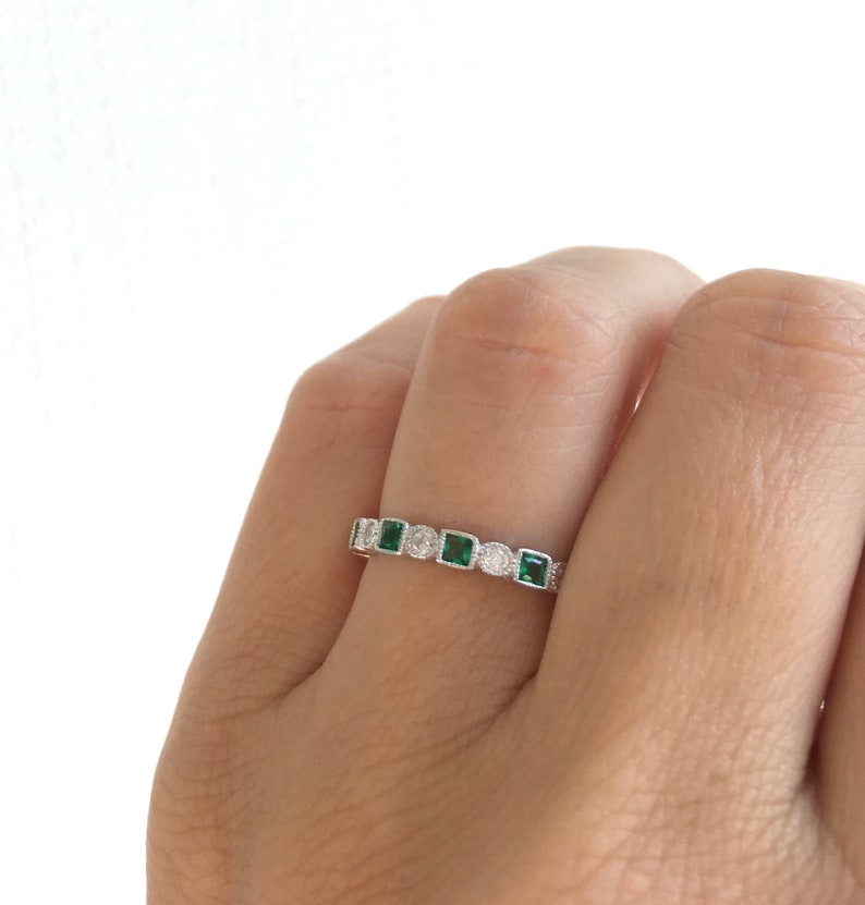 May include: A silver ring with a repeating pattern of square emerald-green stones and round clear stones. The ring is set with small, round accent stones between the larger stones. The ring is on a finger.