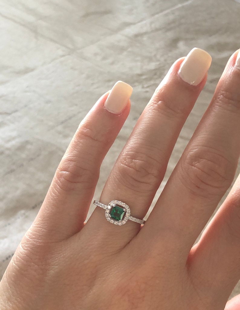 May include: A silver ring with a square emerald green gemstone surrounded by a halo of smaller clear gemstones.