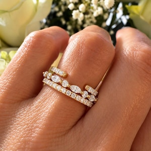 May include: A gold ring featuring a multi-layered design with clear stones. The band includes a row of round pavé stones, a central row alternating between marquise and round stones, and an open-ended top section with pavé stones.