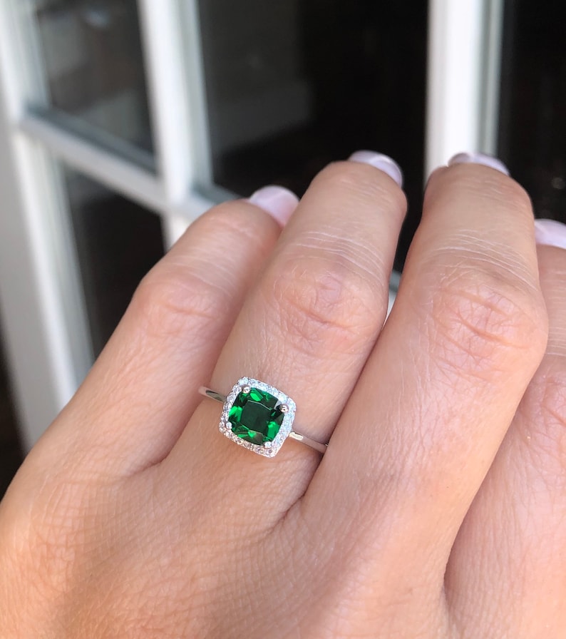 Puede incluir: Un anillo de plata con una piedra preciosa cuadrada verde esmeralda rodeada por un halo de piedras preciosas m&aacute;s peque&ntilde;as y transparentes.