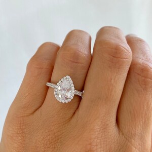 May include: A pear-shaped engagement ring with a halo setting. The ring is set in silver and features a large, clear center stone surrounded by smaller diamonds.