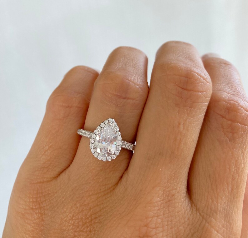 May include: A pear-shaped engagement ring with a halo setting. The ring is set in silver and features a large, clear center stone surrounded by smaller diamonds.