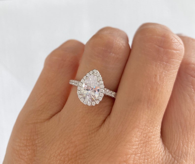 May include: A pear-shaped engagement ring with a halo setting. The ring is made of silver and has a clear center stone.